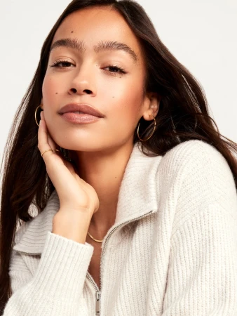 Close-up portrait of a woman with dark hair wearing a white ribbed knit zip-up sweater against a white seamless background.