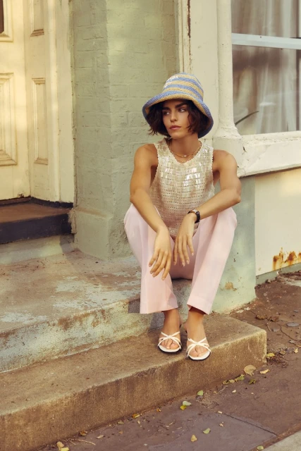 Young woman wearing a striped bucket hat, sequined top, and pink pants is sitting on concrete steps in front of a painted door.