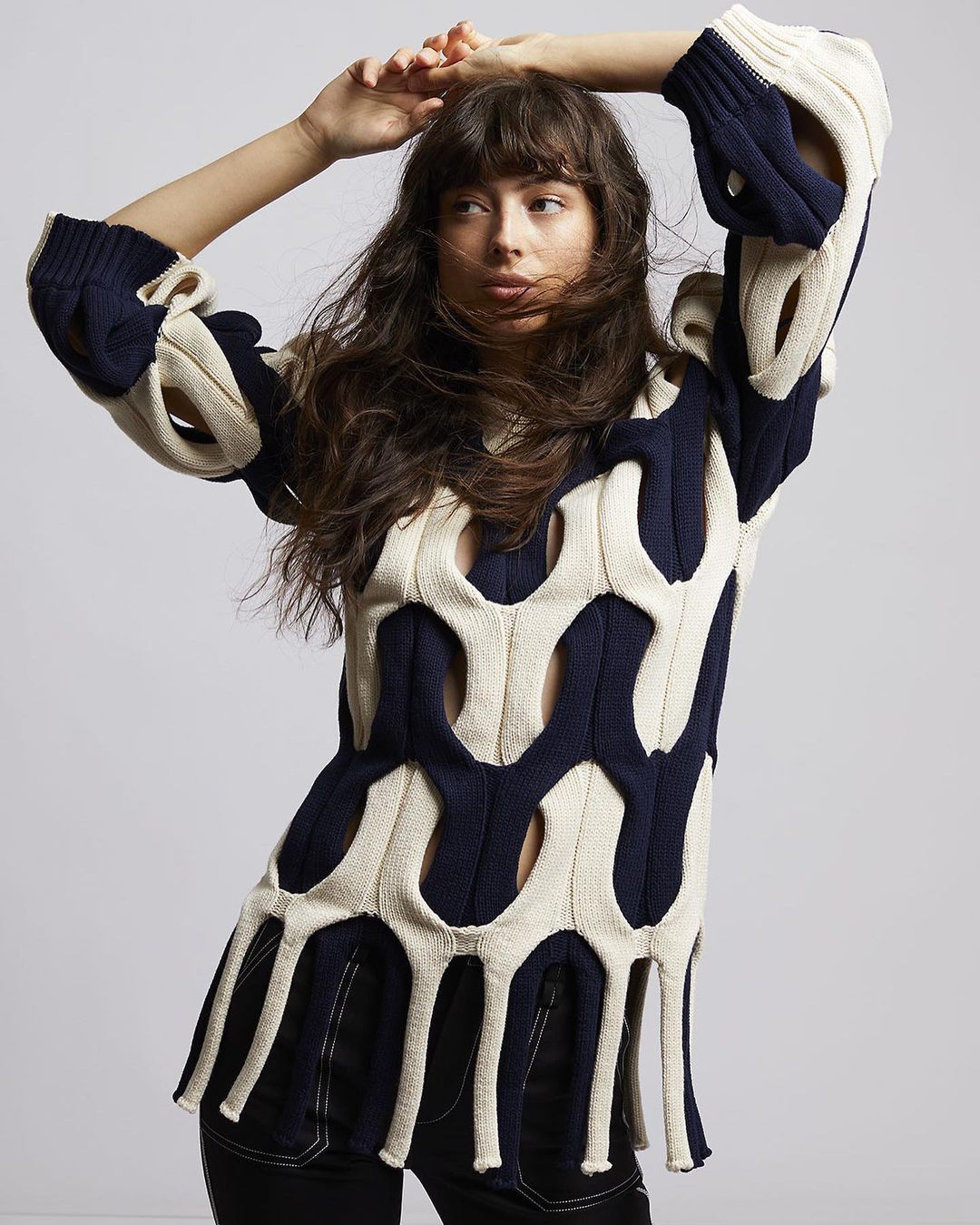 Woman wearing a cut-out cream and navy knit sweater with fringe hem, posing against a plain grey backdrop.
