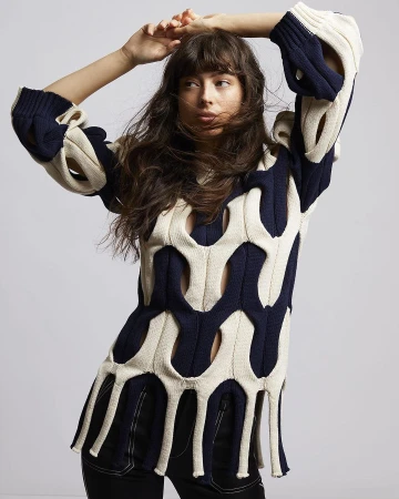 Woman wearing a cut-out cream and navy knit sweater with fringe hem, posing against a plain grey backdrop.