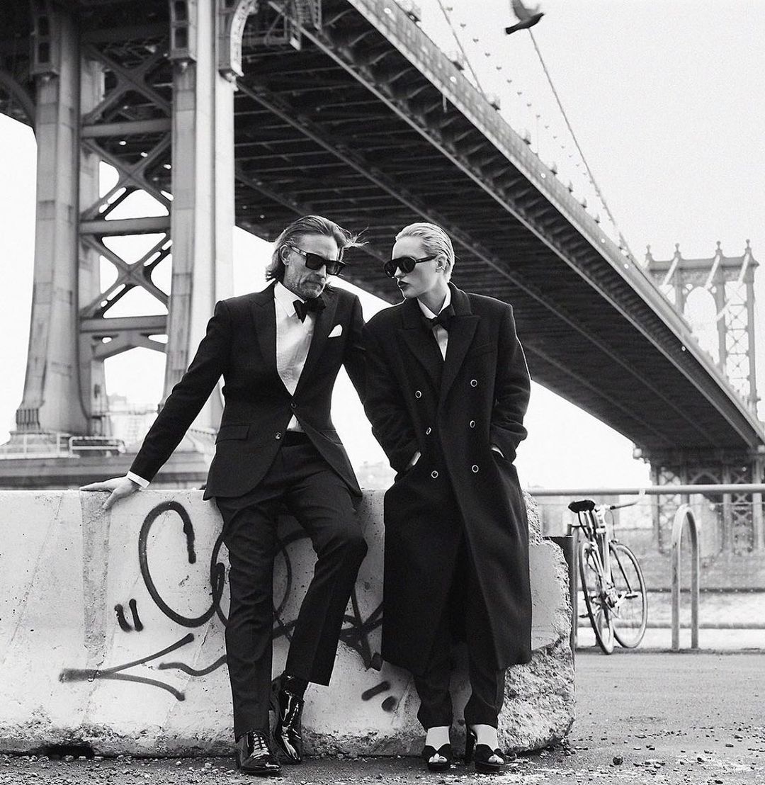 A couple in formal attire pose in front of the Manhattan Bridge; a man in a suit leans against a concrete barrier near a bike.