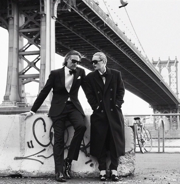 A couple in formal attire pose in front of the Manhattan Bridge; a man in a suit leans against a concrete barrier near a bike.