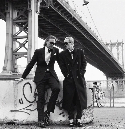 A couple in formal attire pose in front of the Manhattan Bridge; a man in a suit leans against a concrete barrier near a bike.