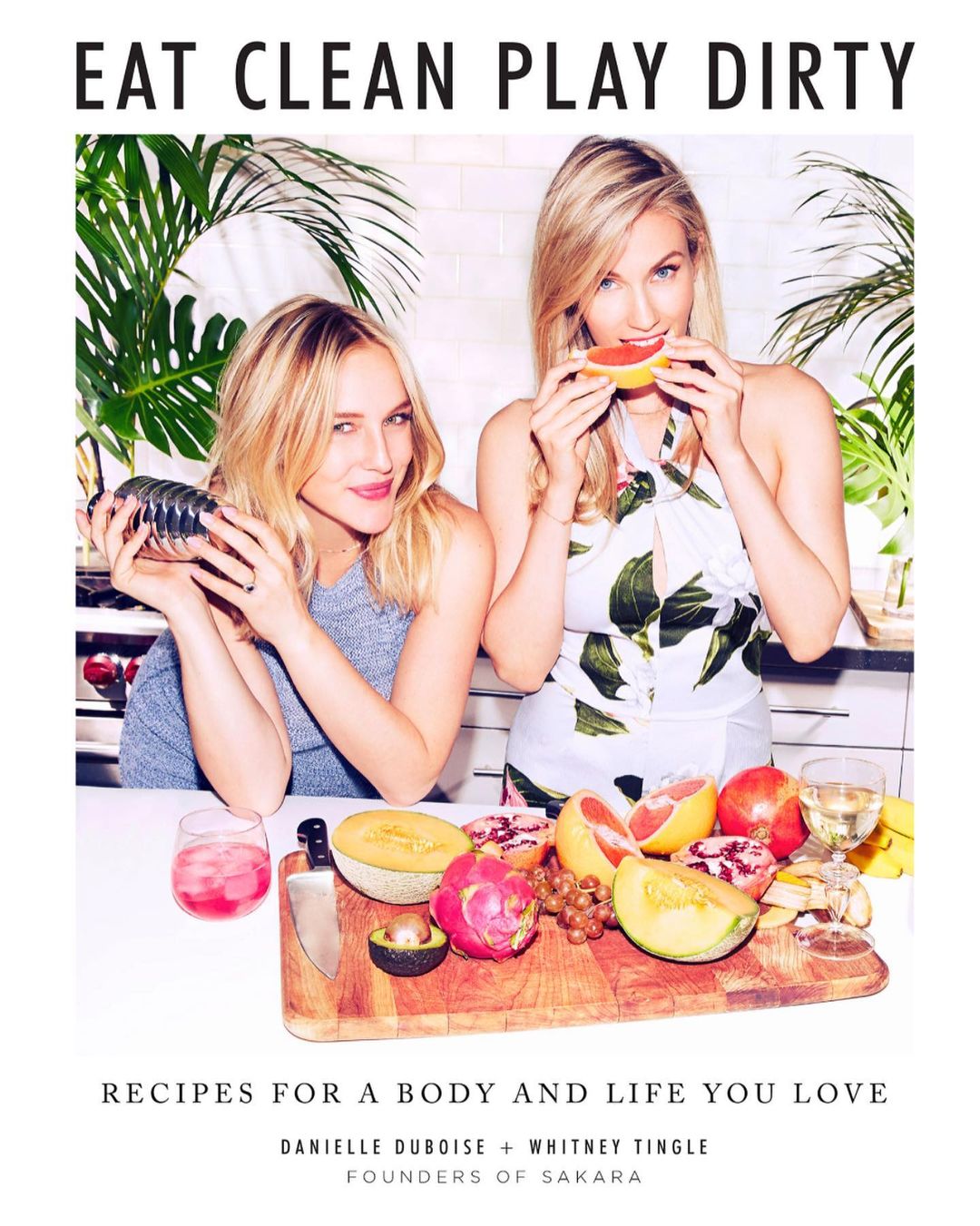 Two blonde women in a bright, indoor kitchen setting with tropical fruit, one holding a shaker, the other eating grapefruit.