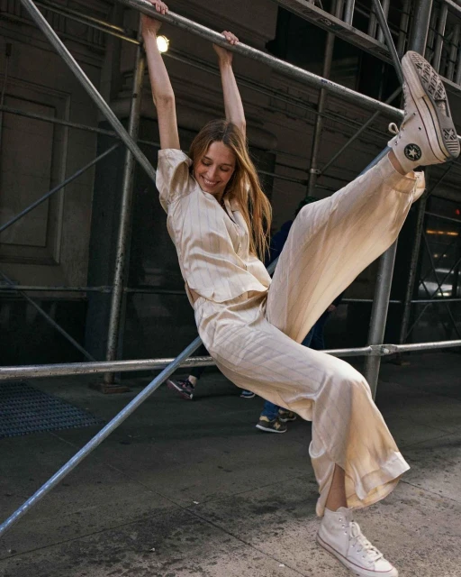 A cheerful woman in a light-colored striped pantsuit and sneakers playfully hangs from metal scaffolding on a city street with other people walking by.