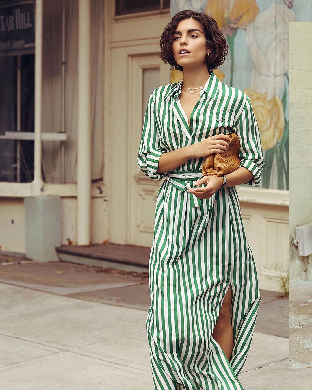 Elegant woman in green-striped dress with clutch poses on city sidewalk. Mid-shot showcases fashion and urban style.