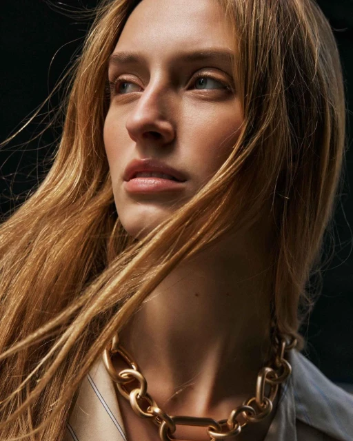 Close-up of a light-skinned adult woman with long blonde hair and a gold chain necklace, posing against a dark background.