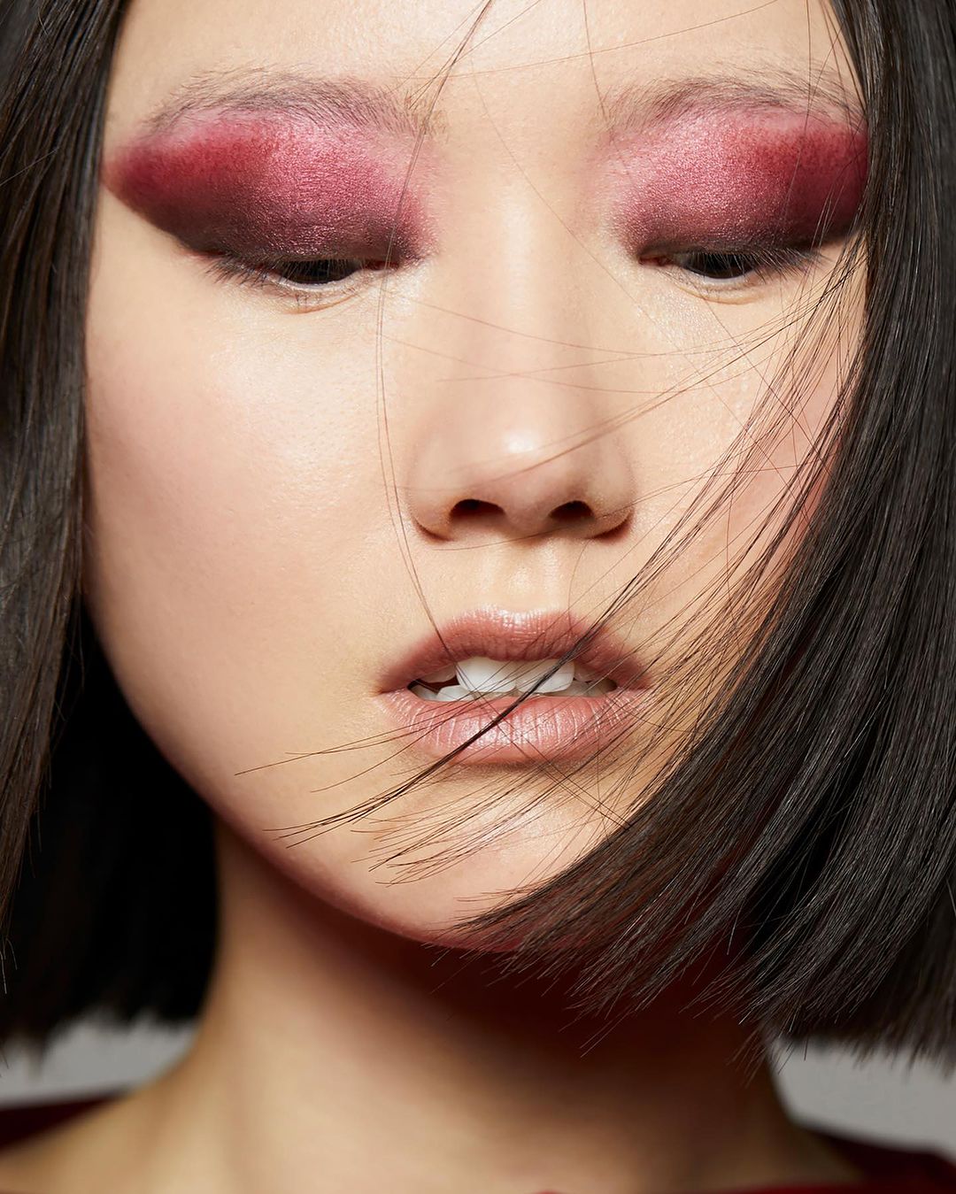 Close-up beauty portrait of an East Asian woman with dramatic dark pink eyeshadow and straight dark hair blowing across her face.