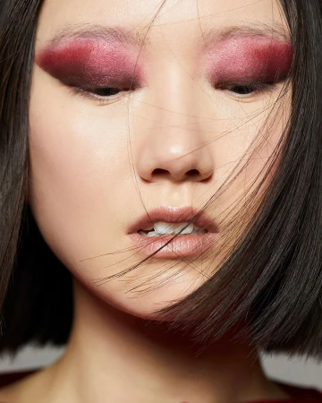 Close-up beauty portrait of an East Asian woman with dramatic dark pink eyeshadow and straight dark hair blowing across her face.