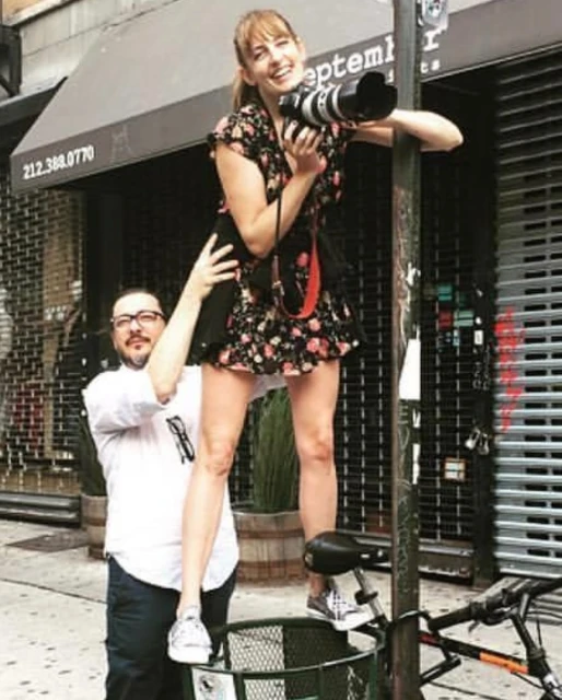 Female photographer stands on a trash can on a city street, aiming a camera while a man steadies her, capturing an urban scene.