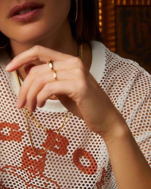 Close-up of an adult female wearing gold hoop earrings and a white fishnet top with gold necklaces and a ring on her finger.