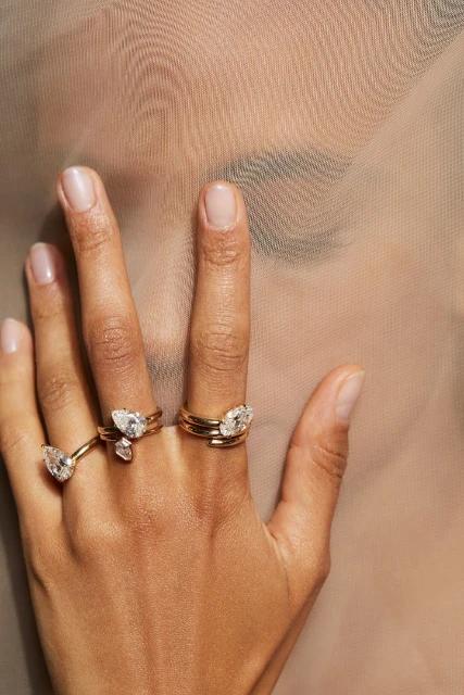 Close-up of a hand with multiple gold rings adorned with pear-shaped diamonds, set against a softly blurred neutral-toned background.