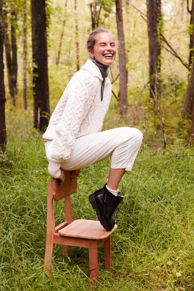Woman in a cable knit sweater and cream pants laughs while sitting on a chair outdoors in a wooded area with dappled sunlight.