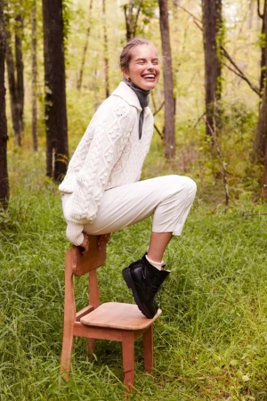 Woman in a cable knit sweater and cream pants laughs while sitting on a chair outdoors in a wooded area with dappled sunlight.