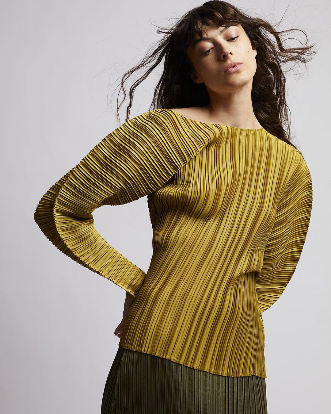 Brunette adult woman poses in a studio, wearing a pleated chartreuse long-sleeve top and a dark green pleated skirt against a gray backdrop.