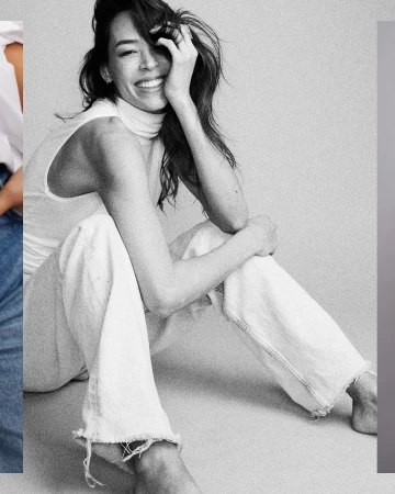 Candid black and white portrait of a smiling woman sitting on the floor wearing a sleeveless turtleneck top and light denim jeans.