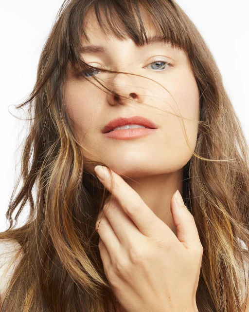 Close-up studio portrait of a young adult woman with long brown hair and bangs touching her face, set against a clean white background.