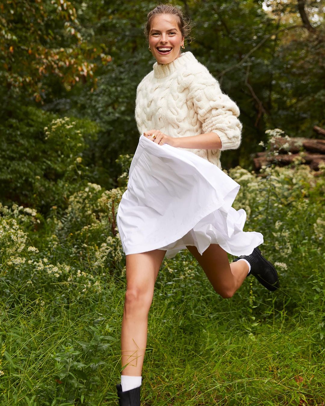 Smiling woman in sweater and skirt running outdoors through a grassy field near trees, showcasing fashion and active lifestyle in nature.