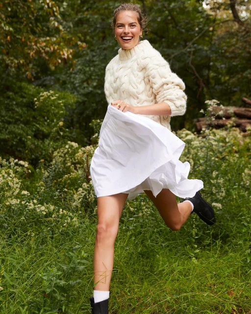 Smiling woman in sweater and skirt running outdoors through a grassy field near trees, showcasing fashion and active lifestyle in nature.