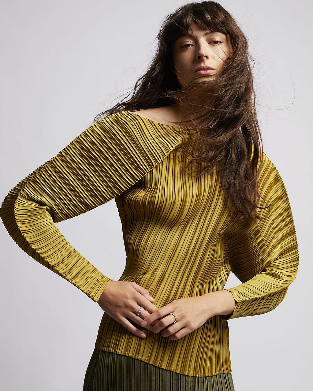 Brunette woman poses in a studio wearing a pleated chartreuse blouse and olive skirt, showcasing fashion and style in soft lighting.