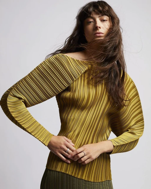 Brunette woman poses in a studio wearing a pleated chartreuse blouse and olive skirt, showcasing fashion and style in soft lighting.
