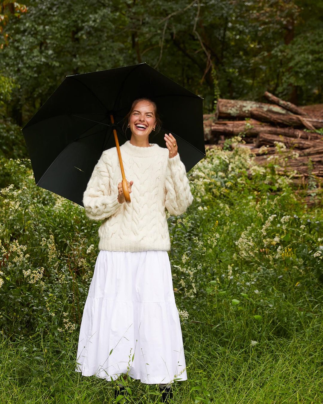A happy adult female stands in a grassy field under an umbrella, wearing a white knit sweater and a long white skirt, laughing.