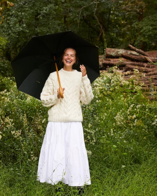 A happy adult female stands in a grassy field under an umbrella, wearing a white knit sweater and a long white skirt, laughing.