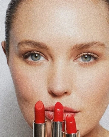 Close-up portrait of a woman holding three red lipsticks in silver tubes near her face against a neutral background.