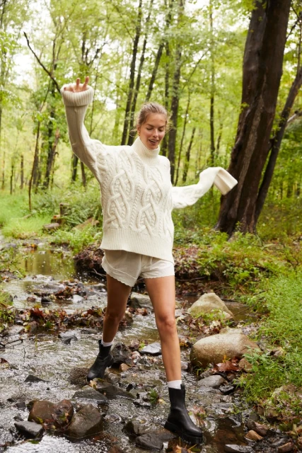 A young adult woman in a white sweater and shorts raises her arms in a forest creek surrounded by green trees and foliage.