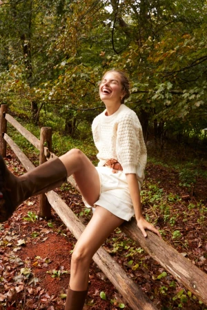 Woman in cream knit sweater and white shorts laughing while seated on a wooden fence outdoors