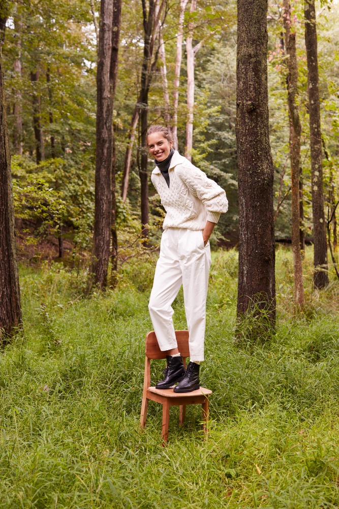 Woman in cream cable-knit sweater and white trousers standing on wooden chair in forest