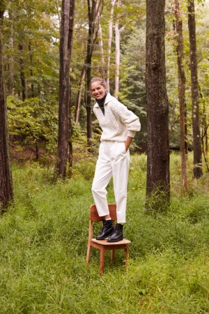Woman in cream cable-knit sweater and white trousers standing on wooden chair in forest