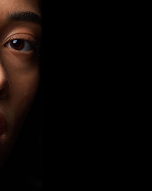 Close-up portrait of a woman's face with fair skin, captured in a dramatic half-light composition against a solid black background.