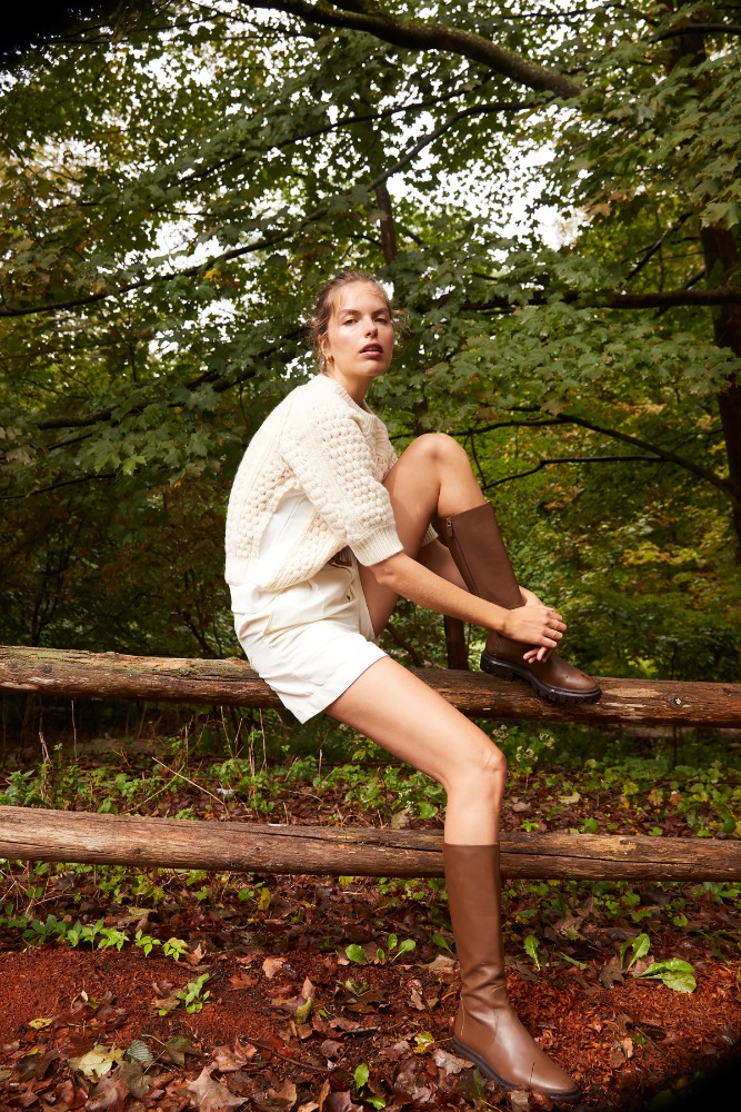 A woman in a cream sweater, white shorts, and brown knee-high boots sits on a wood fence in a wooded outdoor setting.