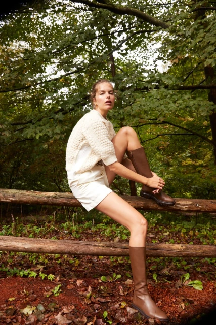 A woman in a cream sweater, white shorts, and brown knee-high boots sits on a wood fence in a wooded outdoor setting.