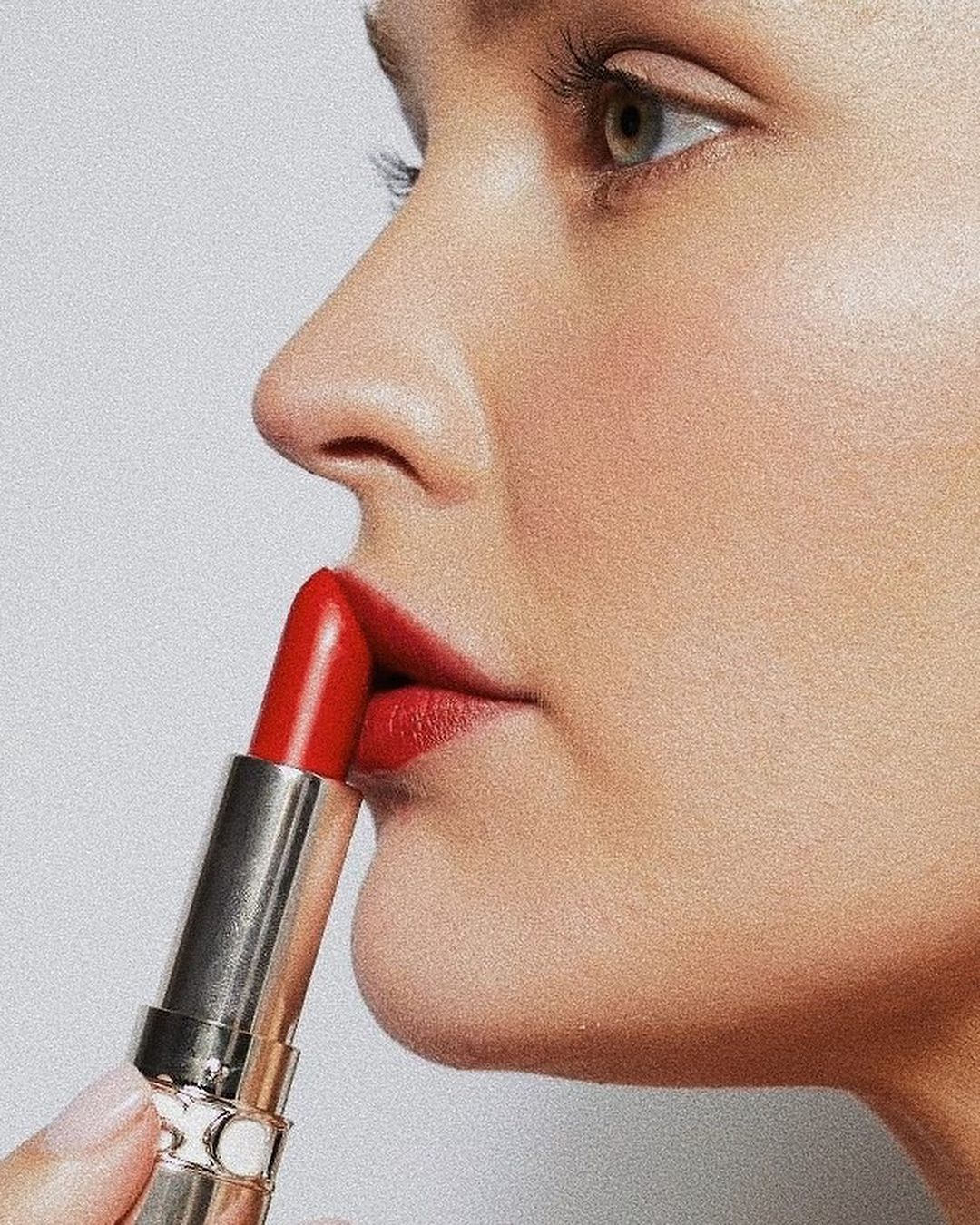 Close-up shot of a fair-skinned adult female applying bright red lipstick, showcasing beauty and makeup in a studio setting against a white backdrop.
