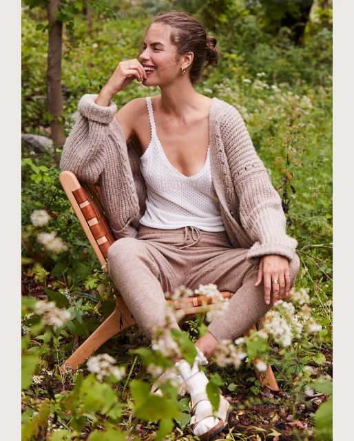 A woman is smiling while sitting on a chair in a garden, wearing a white tank top and beige knit cardigan and matching sweatpants.