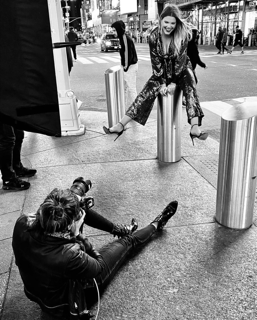 Candid black and white street style photograph of a smiling adult woman in a sequined outfit, seated on a bollard, being photographed.