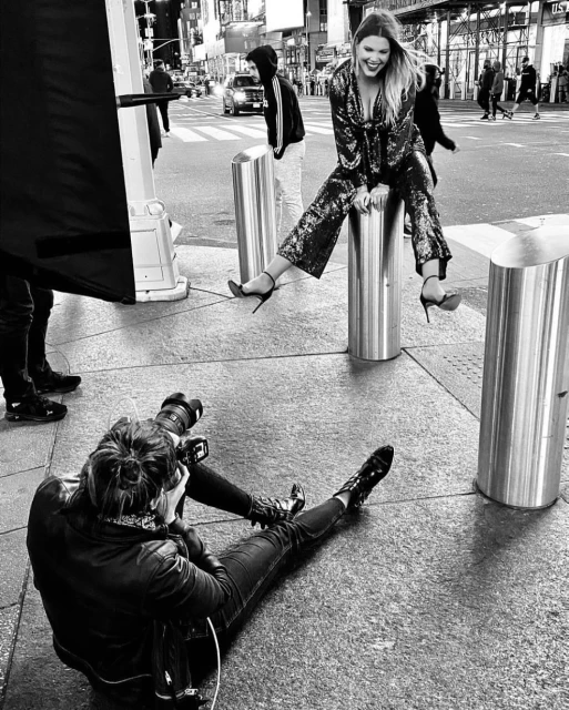 Candid black and white street style photograph of a smiling adult woman in a sequined outfit, seated on a bollard, being photographed.
