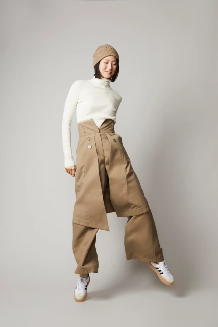 Fashionable young woman in a turtleneck, beige beanie, and khaki pants poses in a studio with a neutral background, showcasing stylish apparel.
