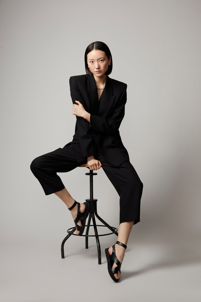 Elegant Asian woman in an oversized black suit and black sandals poses seated on a stool in a neutral studio setting, full body fashion shot.