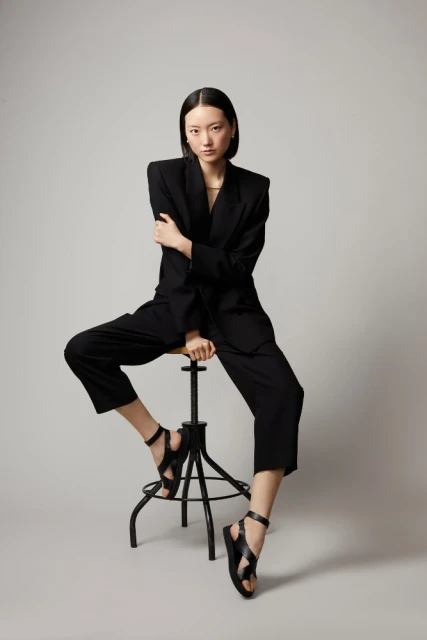 Elegant Asian woman in an oversized black suit and black sandals poses seated on a stool in a neutral studio setting, full body fashion shot.