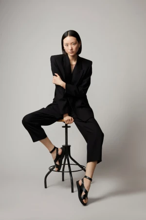 Elegant Asian woman in an oversized black suit and black sandals poses seated on a stool in a neutral studio setting, full body fashion shot.
