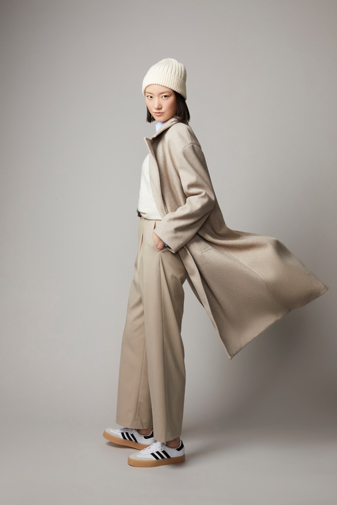 A full shot of a young Asian woman in a beige wool coat, matching slacks, and a cream-colored beanie posing in a photo studio.