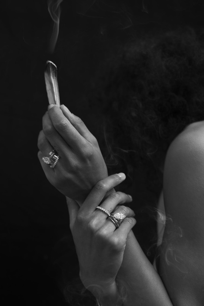 Close-up monochrome portrait of hands holding a smoking sage bundle adorned with diamond rings.