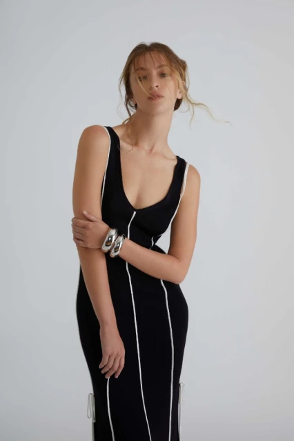 A slender adult woman in a black dress with white trim stands in a bright studio with minimal makeup and hair up in a casual style.