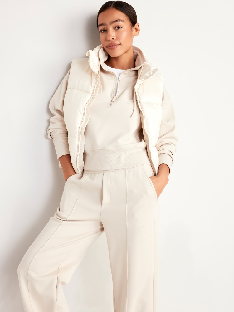 Young woman in a cream-colored sweatsuit and a puffer vest stands against a neutral background in a fashion-style portrait.