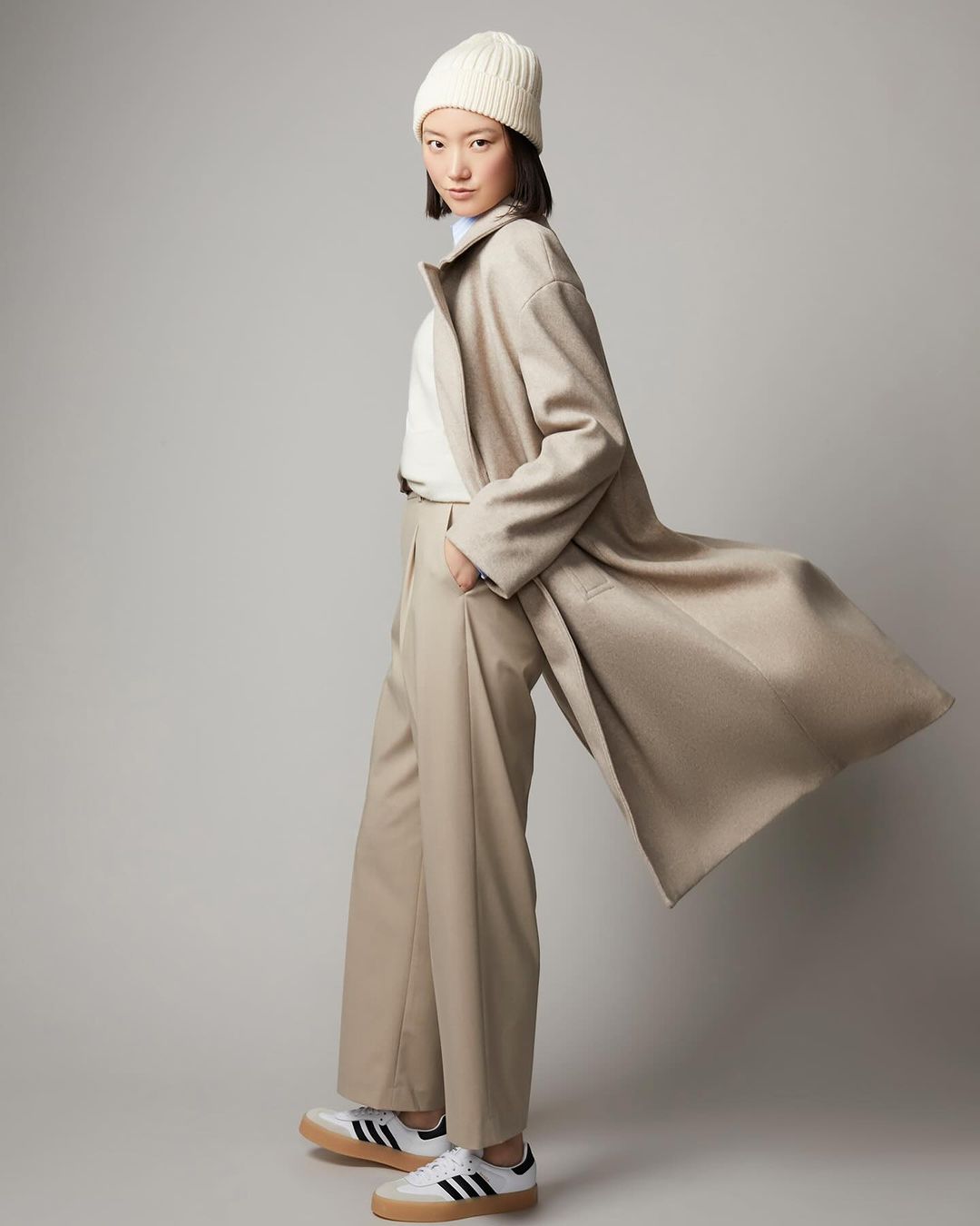 An Asian woman stands in a photo studio wearing a beige wool coat, matching trousers, a cream-colored beanie, and Adidas Samba sneakers.