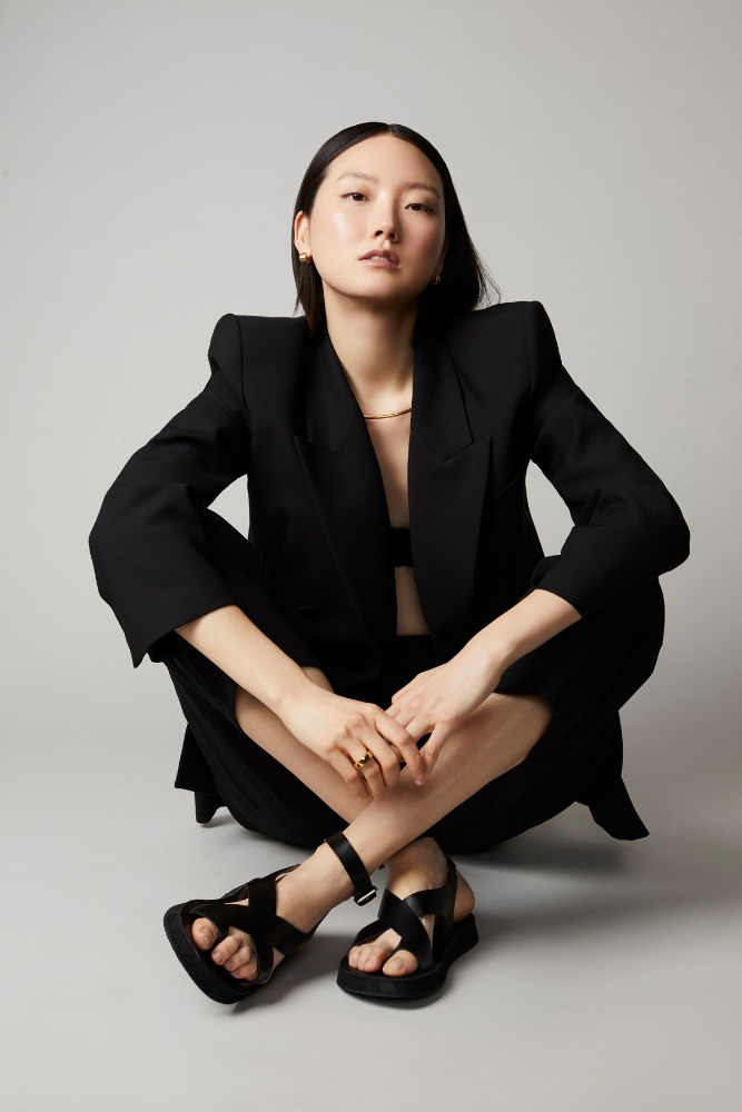 East Asian woman seated on the floor wearing an oversized black blazer and black sandals against a grey studio backdrop.