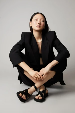 East Asian woman seated on the floor wearing an oversized black blazer and black sandals against a grey studio backdrop.
